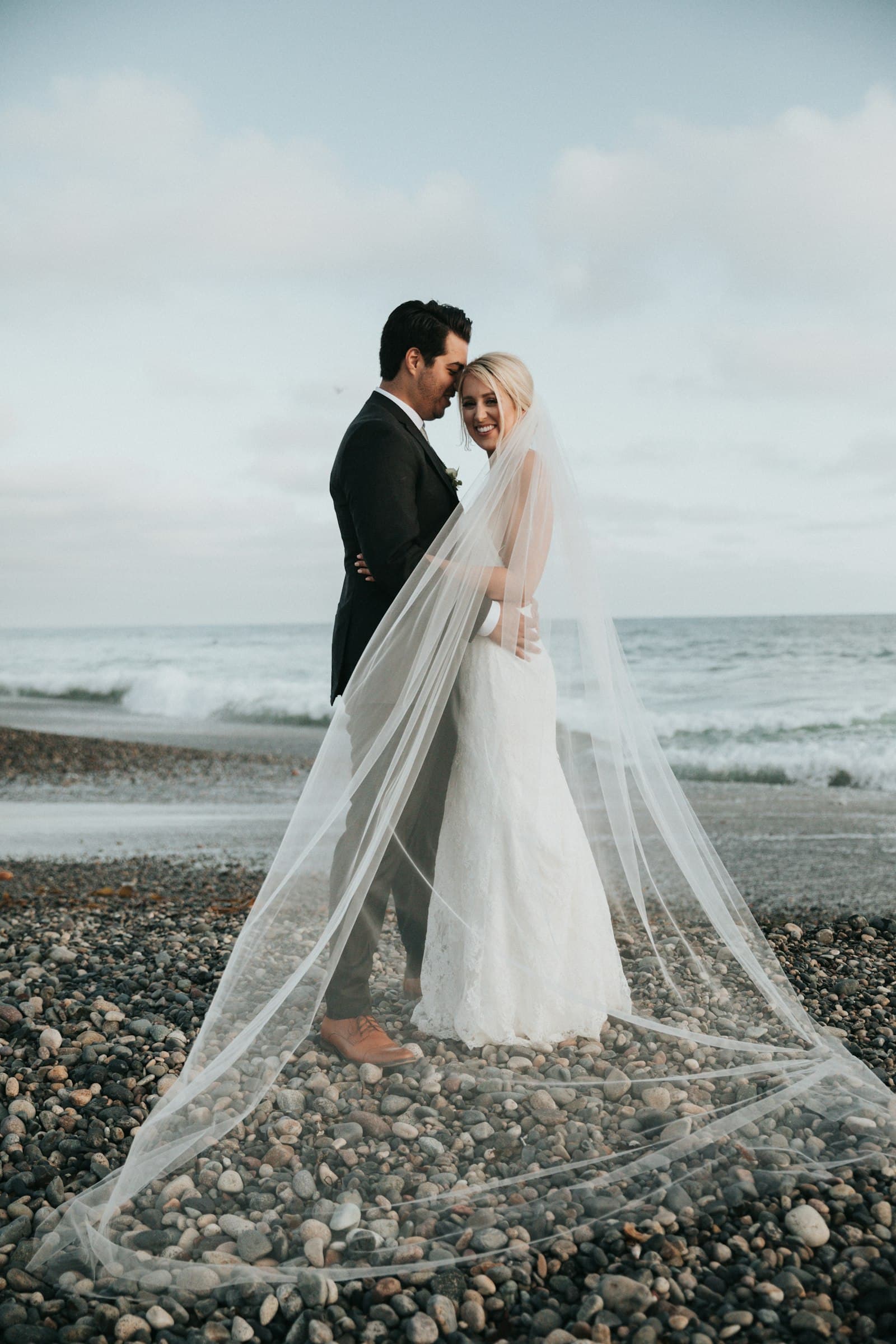 Couple embracing on a pebble beach under a long veil